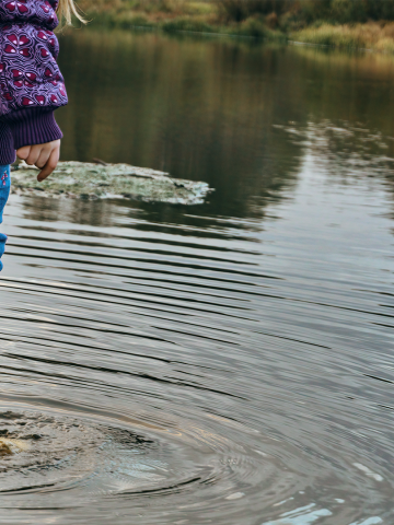 A child in yellow rubber boots stands in the water, blue jeans and a purple jacket, with her feet under water in a flooded street.
