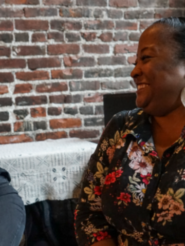 Two women sitting and smiling at each other in a warmly lit space with a brick wall background. One woman is wearing a black blouse, and the other is in a floral print top with large earrings. 