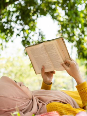 Photo of a young Muslim woman reading a book on a red checkered blanket with a picnic basket with yellow sunflowers nearby. She is surrounded by green trees.