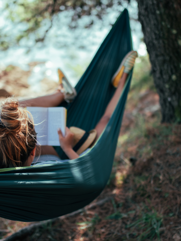 Photo of a woman reading a book in a hammock in the forest nearby a river