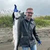 Headshot of Joe Sonnier, director of programs and grant operations at Alaska Community Foundation, standing outdoors on a sandy shoreline holding a large fish with one hand. He is wearing a dark jacket, blue gloves, and jeans, with tall grass and a cloudy sky in the background.