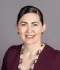 Headshot of Nichole June Maher smiling, wearing a burgundy top, gold earrings and a beaded necklace, against a plain gray background.