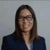 Headshot of Jodi Nishioka, an Asian woman with shoulder-length dark hair, wearing glasses and a navy blazer over a patterned shirt, smiling in front of a plain gray background.