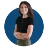 Headshot of Audrey against a blue background. Audrey wears a black short-sleeve top, olive-green high-waist pants, a pendant necklace, and teal drop earrings.