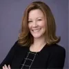 Headshot of Traci McGlathery, STCU’s Here for Good, smiling with arms crossed, wearing a black blazer over a patterned top, shoulder-length light brown hair, photographed against a soft purple backdrop.