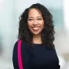 Headshot of Angela Jones, Gates Foundation, smiling at the camera. She has shoulder-length curly dark hair and wears a navy dress with a bright pink stripe on the sleeve, photographed against a softly blurred indoor background.