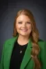 Headshot of Kathleen Chick, smiling at the camera, wearing a green blazer over a black top with a small gold pendant necklace, against a dark gray studio background.