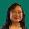 Headshot of Alice Ito, smiling, with shoulder-length dark hair and round glasses, wearing a dark patterned top against a pine green background.