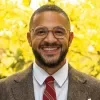 Headshot of Jesse Beason, smiling at the camera, wearing round glasses, a brown blazer, white shirt, and red striped tie, photographed against a softly blurred background of bright yellow leaves.