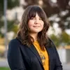 Headshot of Lauren Waudé, standing outdoors with a calm expression. She has long dark brown hair with bangs and wears a navy blazer over a mustard top with a geometric pendant necklace, against a softly blurred background.