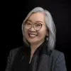 Headshot of Marie Kurose, smiling, with shoulder-length silver-gray hair and red-framed glasses, wearing a dark blazer and scarf against a black studio background.