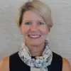 Headshot of Theresa Erstad McLeod, smiling at the camera, wearing a sleeveless black top and a light patterned scarf, with short blonde hair against a neutral textured background.