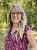 Photo of Jean Chiappisi Oregon School Board Association smiling outdoors, with long blonde hair and glasses, wearing a burgundy floral blouse against a softly blurred green wooded background.