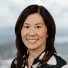 Headshot of Jill Nishi, smiling, with shoulder-length dark hair, wearing a black-and-white patterned top against a softly blurred outdoor background.