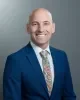 Headshot of Spencer Lewis from Oregon School Board Association smiling with a shaved head, wearing a blue suit jacket, white shirt and colorful floral tie against a gray studio background.