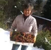Photo of Carmen Davis smiling outdoors in a snowy garden, holding a box of freshly harvested carrots with trees and a deck in the background.