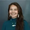 Headshot of Emily Brockman, smiling, with long brown hair, wearing white earrings, a teal sweater and a fish necklace against a dark gray background.