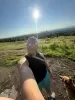 Photo of Janie Caq’ar Ferguson taking a sunny outdoor selfie on a hillside trail, wearing sunglasses and a headband, with a dog beside her and a wide view of trees, water and blue sky in the background.