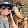 Photo of Jen Cross smiling outdoors, wearing sunglasses and a blue Girdwood hat, with another person seated beside her on a sunny hiking trail surrounded by shrubs.