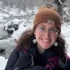 Photo of Shoshi Bieler smiling outdoors in winter, wearing glasses and a brown beanie, with a snowy creek and trees in the background.