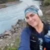 Photo of Sydney Copley taking a selfie beside a rocky river, wearing headphones, a blue headband and a hiking vest with a water bottle.