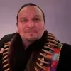 Headshot of George Farrel looking at the camera with a slight smile, wearing a dark shirt and a multicolored scarf or shawl against a plain purple background.