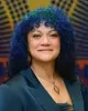 Headshot of Meriah Gille, looking at the camera with a slight smile, with curly blue hair, wearing a dark teal blazer and geometric earrings and necklace against a colorful patterned background.