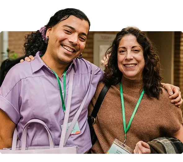 Photograph of two Philanthropy Northwest members smiling. On the left, a person with medium-brown skin and long curly dark hair pulled back wears a lavender shirt and matching bag, with event badges and a green lanyard. On the right, a light-skinned person with long wavy brown hair wears a brown knit top and a green lanyard, holding a coat. Both convey friendliness and connection.