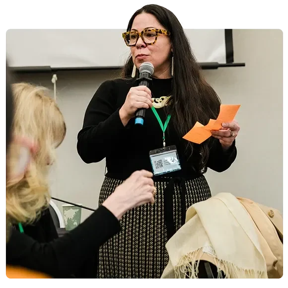 Photograph of Philanthropy Northwest member Se-ah-dom holding a microphone speaking to a group at an event. Se-ah-dom has long dark hair, wears tortoiseshell glasses, a black top, and a patterned skirt with a green lanyard and event badge. Another attendee with light hair is partially visible in the foreground, listening. The scene conveys active participation and engagement.