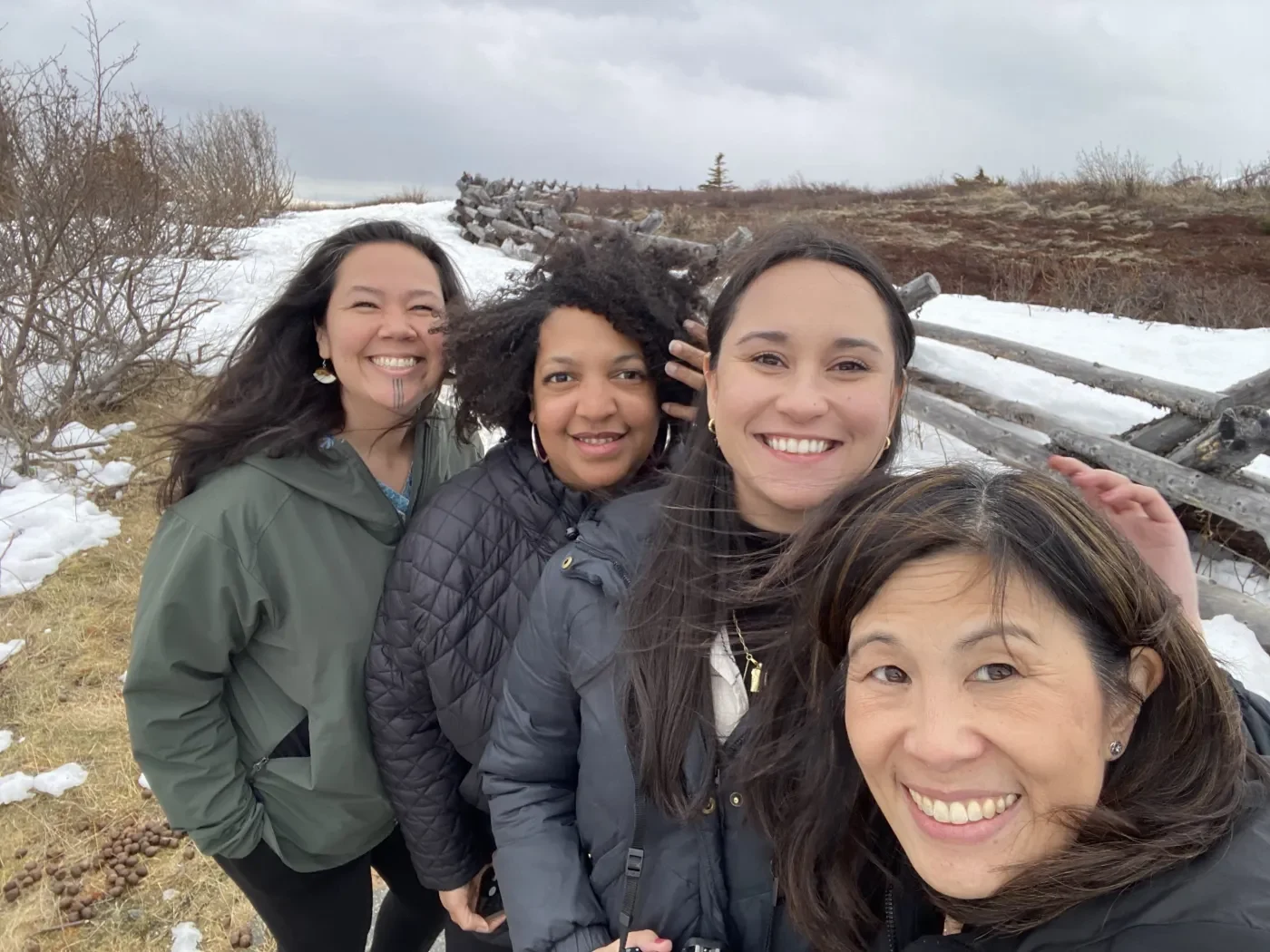 Photo of four people smiling together outdoors in a snowy Alaska landscape, with windblown hair, bare shrubs and a wooden fence behind them.