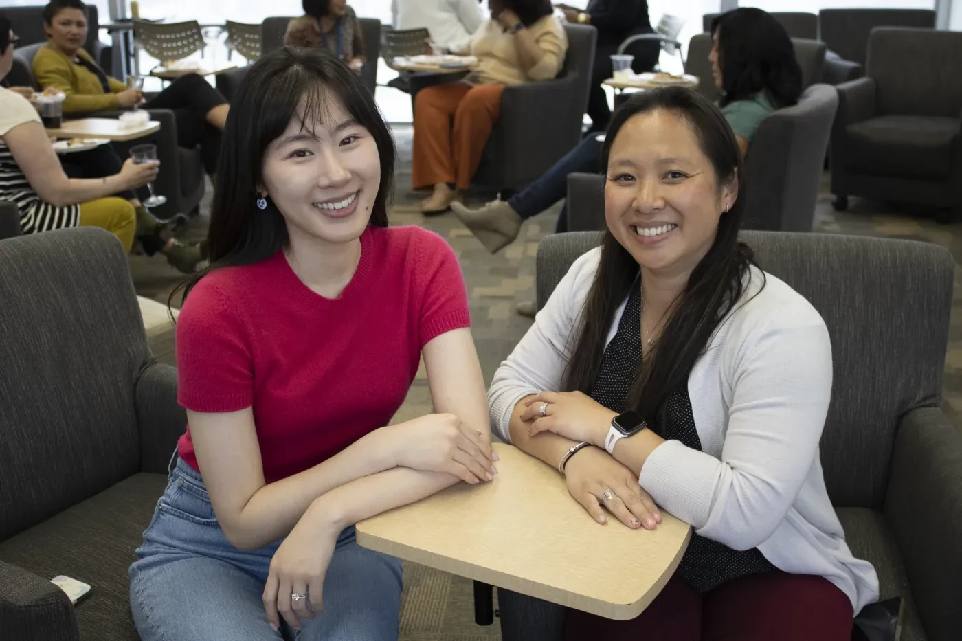 Photo of two people smiling at a small table during an indoor gathering, with other attendees seated and talking in the background.