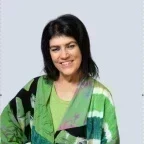 Headshot of a woman with medium-length black hair wearing a green shirt and a multicolored green and black patterned jacket, smiling with her hands on her hips against a light gray background.