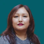 Headshot of Dana Arviso, looking at the camera with a neutral expression, with long auburn-brown hair, wearing dark beaded earrings and a gray sweater against a dark ocean-blue background.