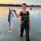 Photo of Emily Brockman standing in shallow water at sunset, smiling while holding up a large fish, with seabirds flying and people wading in the background.