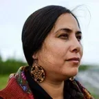 Headshot of Enei Begaye (she/they), a member of the Diné (Navajo) and Tohono O’odham Nations. She faces left in profile with a thoughtful expression, wearing large circular earrings, a dark headscarf, and a patterned scarf. The background is softly blurred, suggesting an outdoor setting.