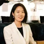 Headshot of Grace Choi with shoulder-length dark hair wearing a light blazer over a black top, seated on a sofa in a modern office with round pendant lights and rows of chairs softly blurred in the background.