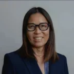 Headshot of Jodi Nishioka, an Asian woman with shoulder-length dark hair, wearing glasses and a navy blazer over a patterned shirt, smiling in front of a plain gray background.