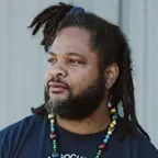 Headshot of Jonathan "JT" Jones-Thomas with long dreadlocks and a beard looking off to the side. He wears a navy T-shirt with “NAACP” printed on it and a beaded necklace with a pendant, standing against a neutral outdoor background.
