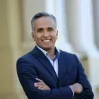 Headshot of Karthick Ramakrishnan, smiling with arms crossed, wearing a navy suit jacket over a light blue shirt in front of softly blurred columns.