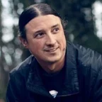 Headshot of Matt Remle, a member of the Hunkpapa Lakota Nation. He looks slightly to the side with a relaxed, thoughtful expression, wearing a dark jacket. The background is softly blurred greenery, giving the portrait a natural, outdoor feel.