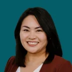 Headshot of Meredith Higashi, smiling, with shoulder-length black hair, wearing a rust-colored blazer over a white top against a dark ocean-blue background.
