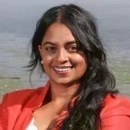 Headshot of Sudha Nandagopal, smiling outdoors, with long dark hair swept to one side, wearing a coral blazer over a white top, with water softly blurred in the background.